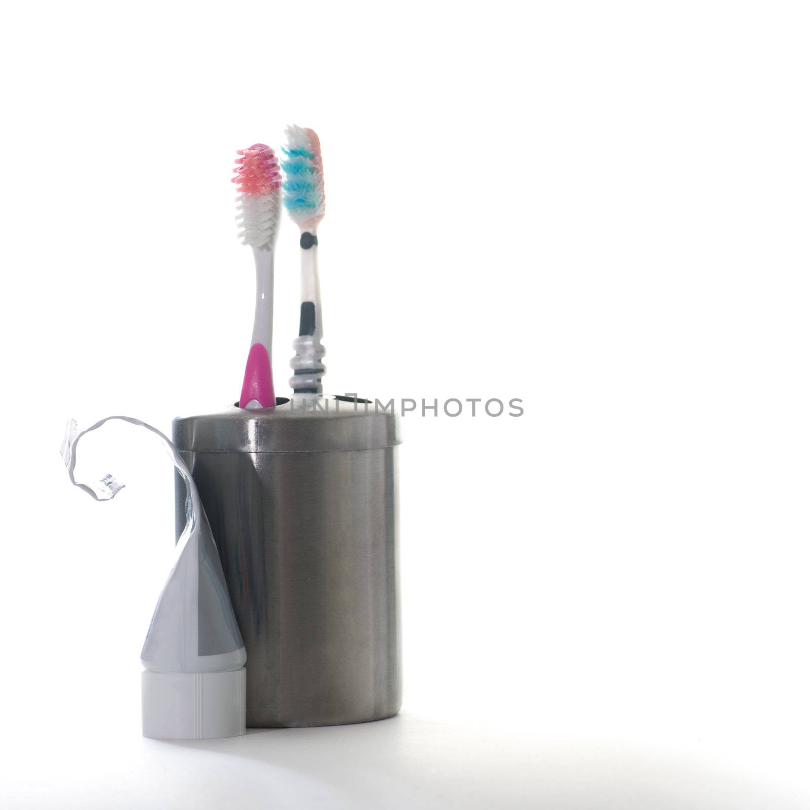 Two toothbrushes in a silver holder, with a half used tube of toothpaste, the end rolled up, isolated on white