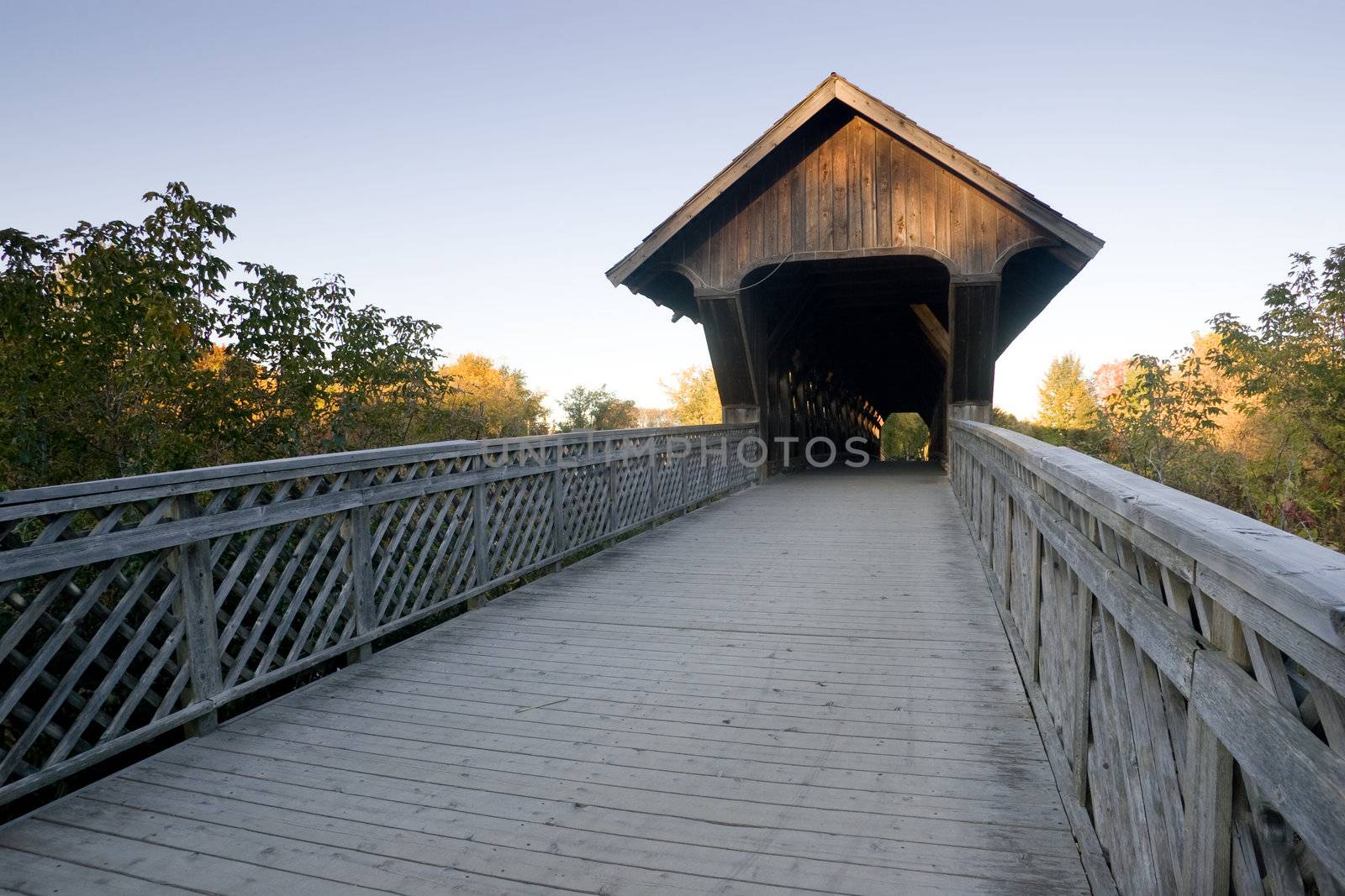 A wide angle view of a covered walking bridge, located in Guelph, Ontario, Canada, with a blue sky background and surrounding fall colors.