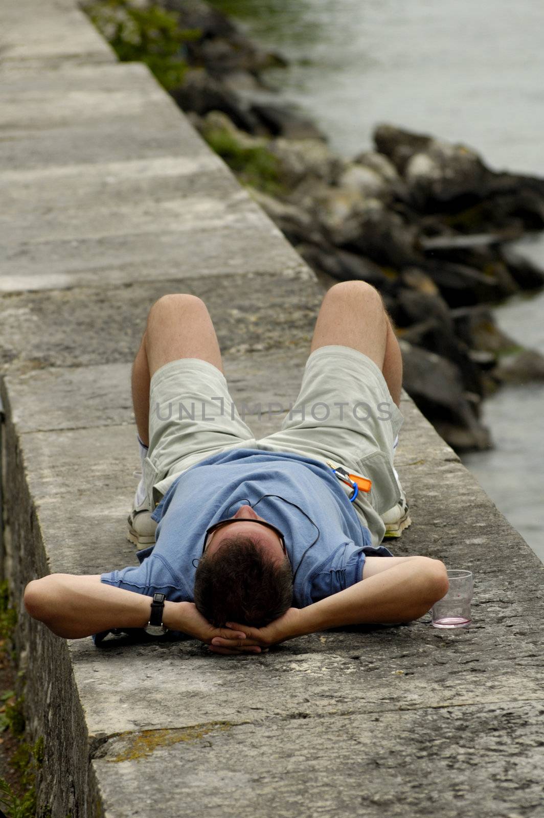 A lone man lies beside a lake, relaxing, with an empty wineglass beside him.