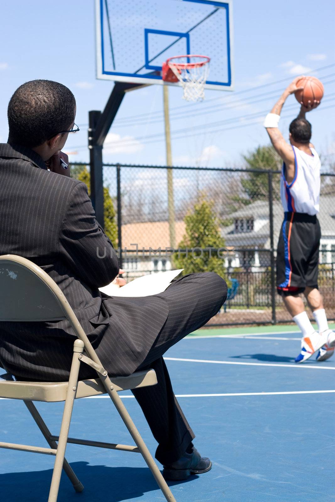 A basketball coach in a business suit observing a player on the team.   He could be also be recruiter trying to scout him.