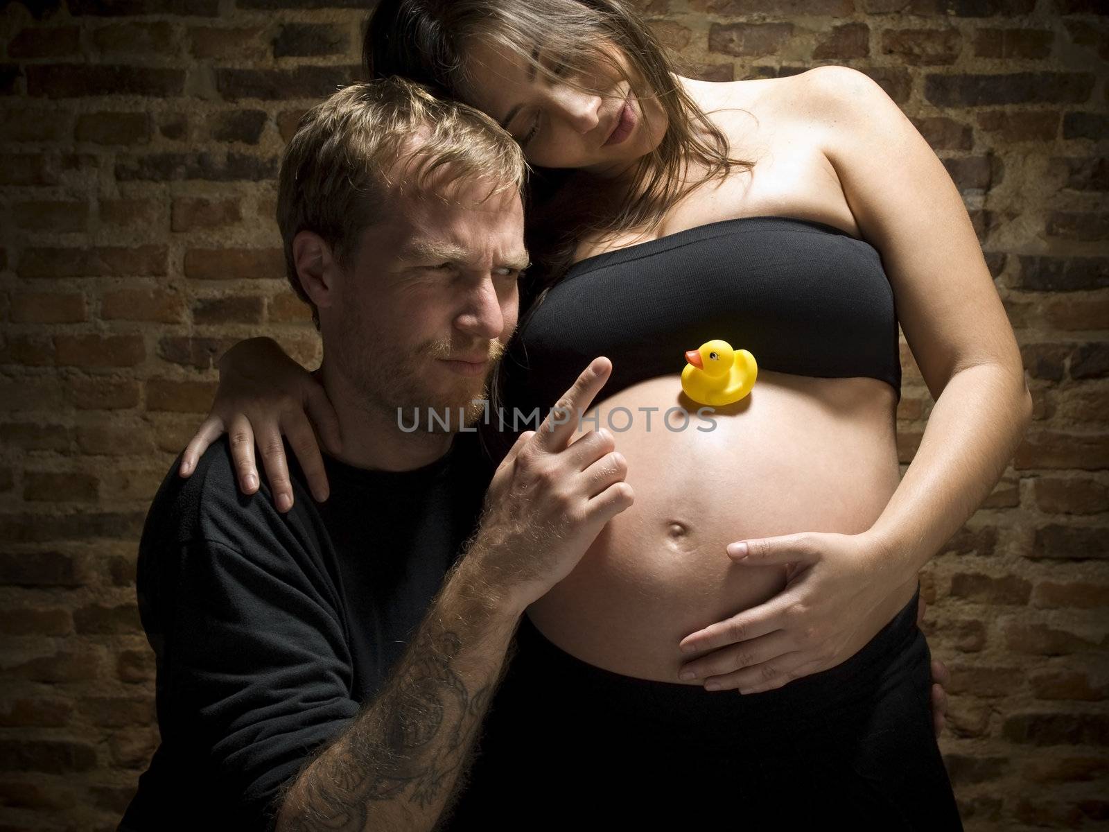 A man pointing his finger at the rubber duck over his wife's pregnant belly.