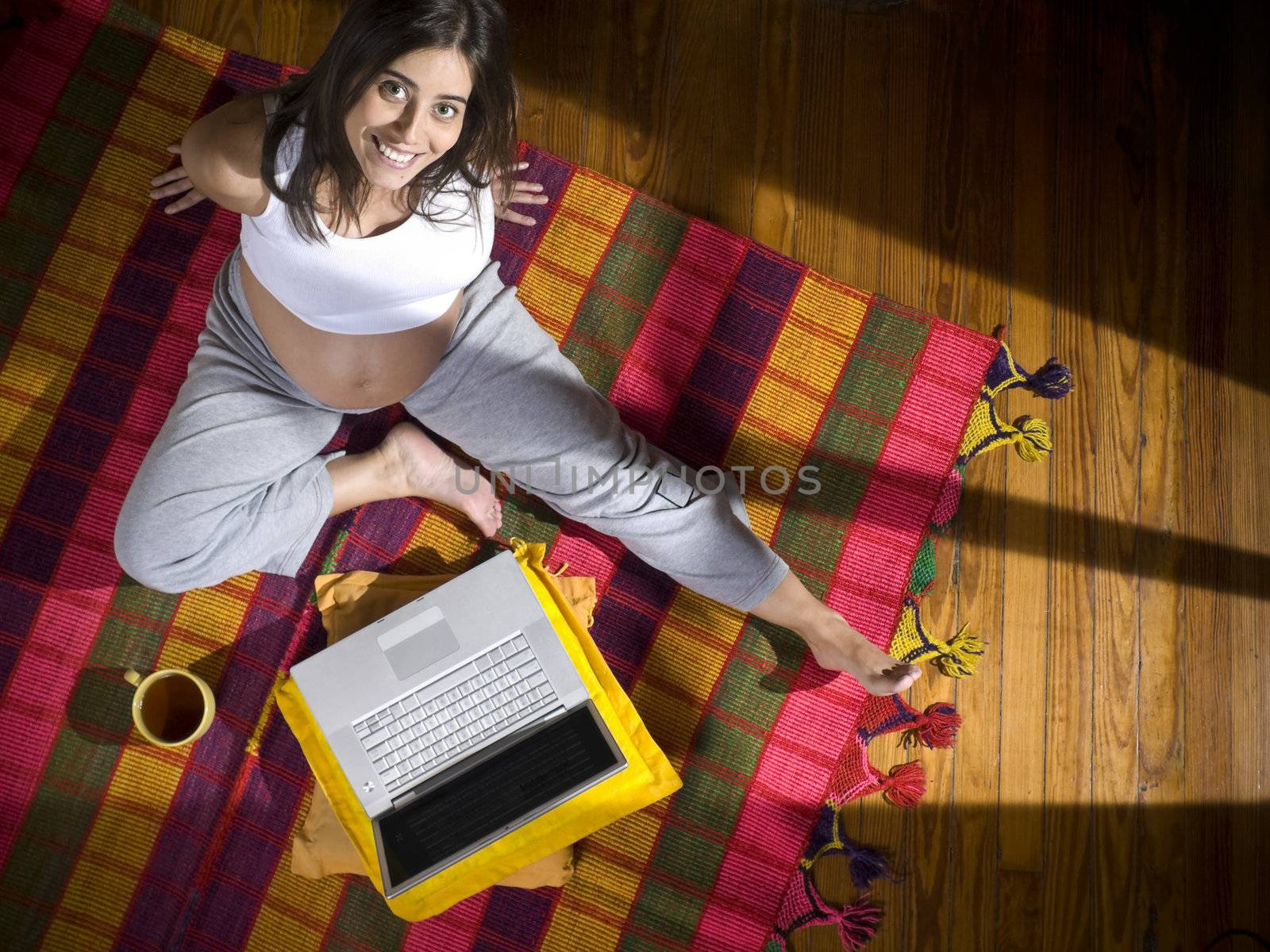 Top view of a young pregnant woman seated on a colorful rug with a laptop computer and a cup of tea.