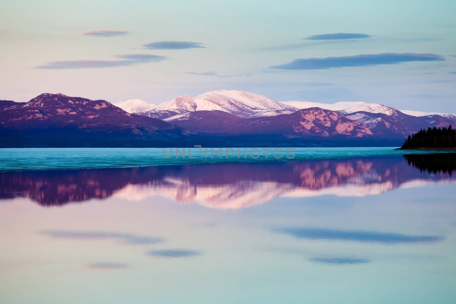 The evening before ice-break at Lake Laberge, Yukon Territory, Canada: reflection of snow-covered mountains on calm open water surface of still largely ice-covered lake.