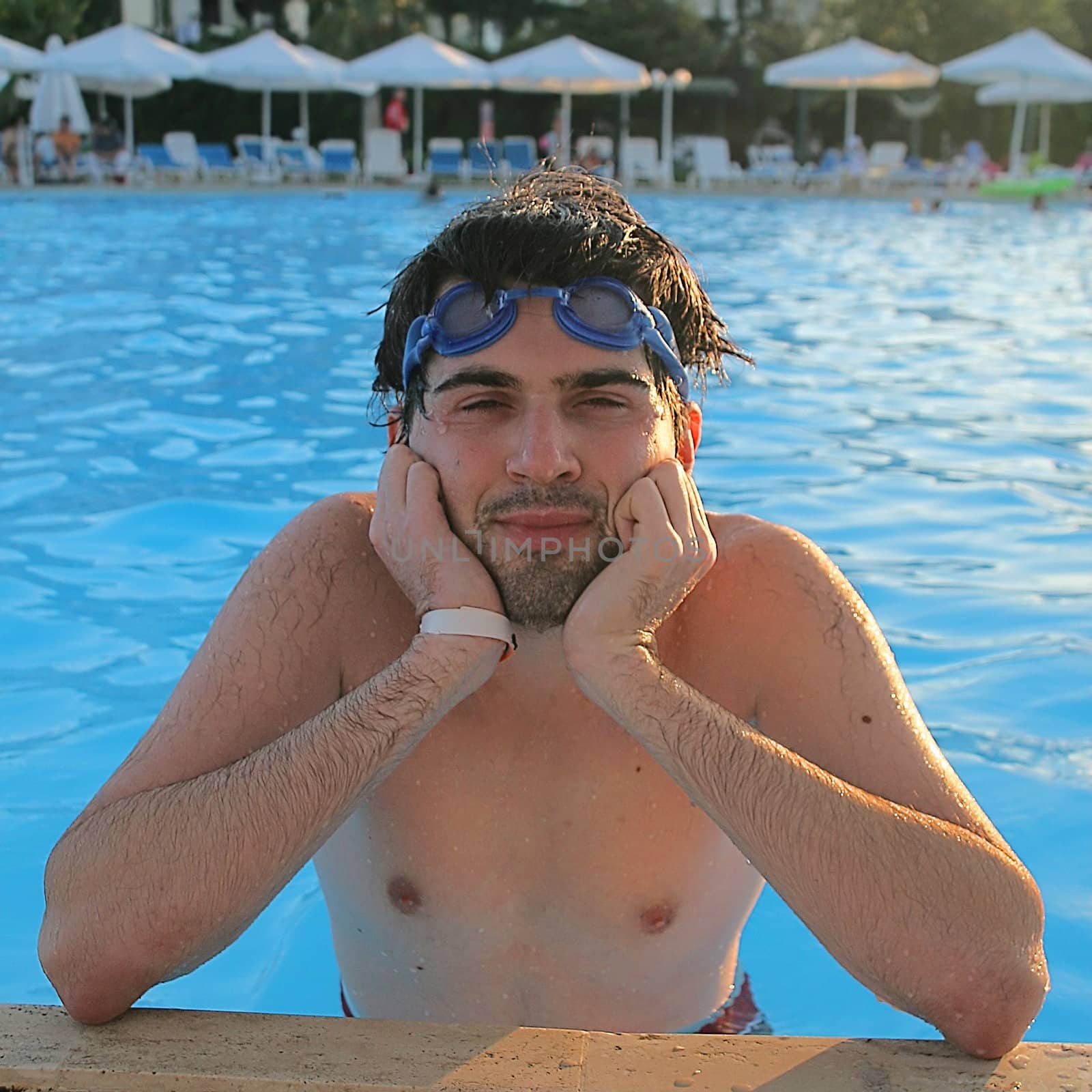 Young man wearing goggles is sitting in the swimming pool leaning to its nosing