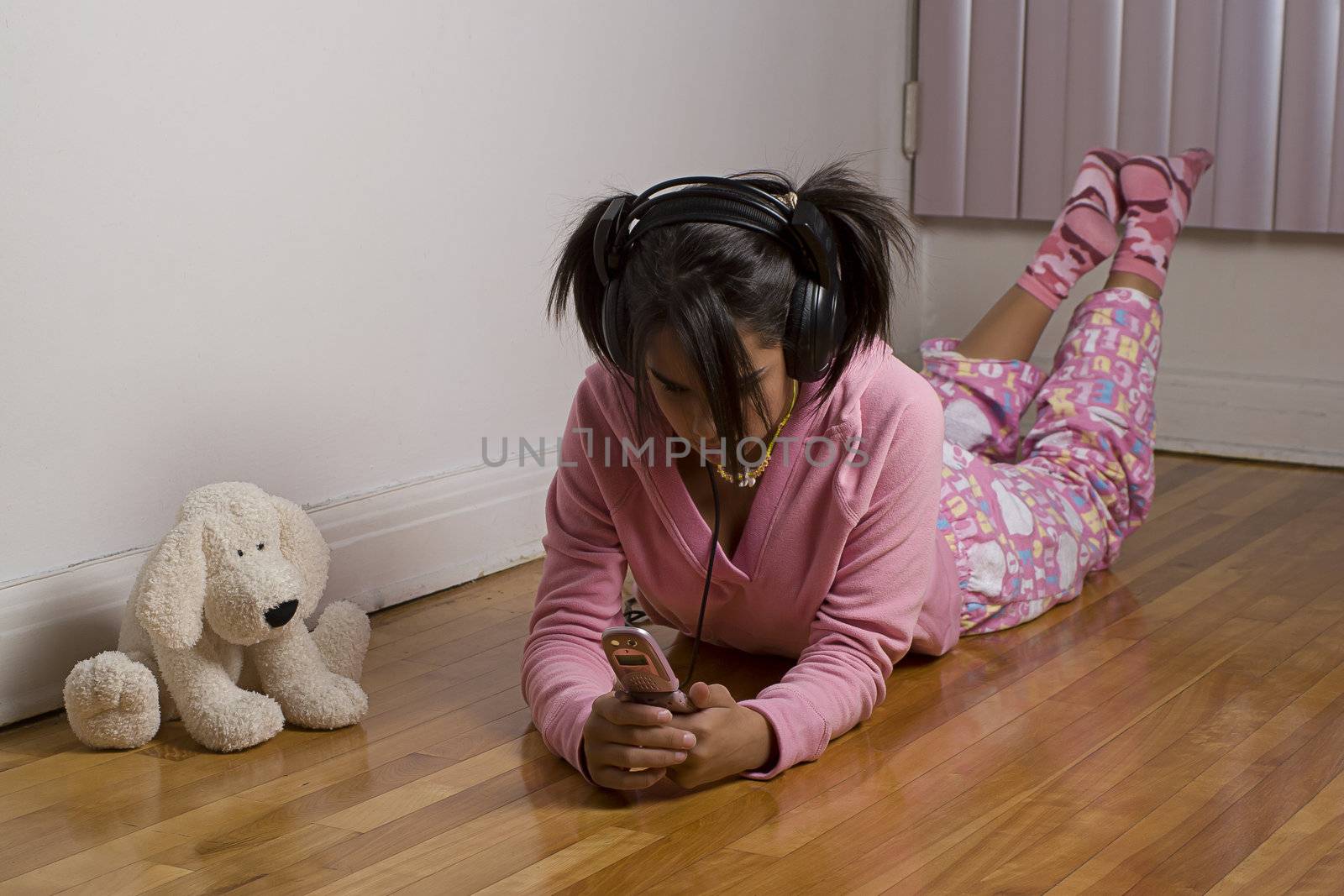 Teenage gir wearing a pink pajama lying on the floor, listing to music and texting on a pink cellphone