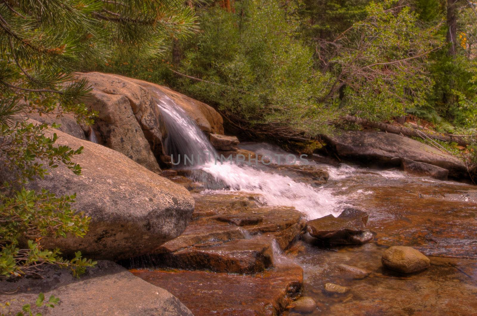 Small single waterfall cascading down mountain boulders into shallow water with forest frame in HDR high dynamic range