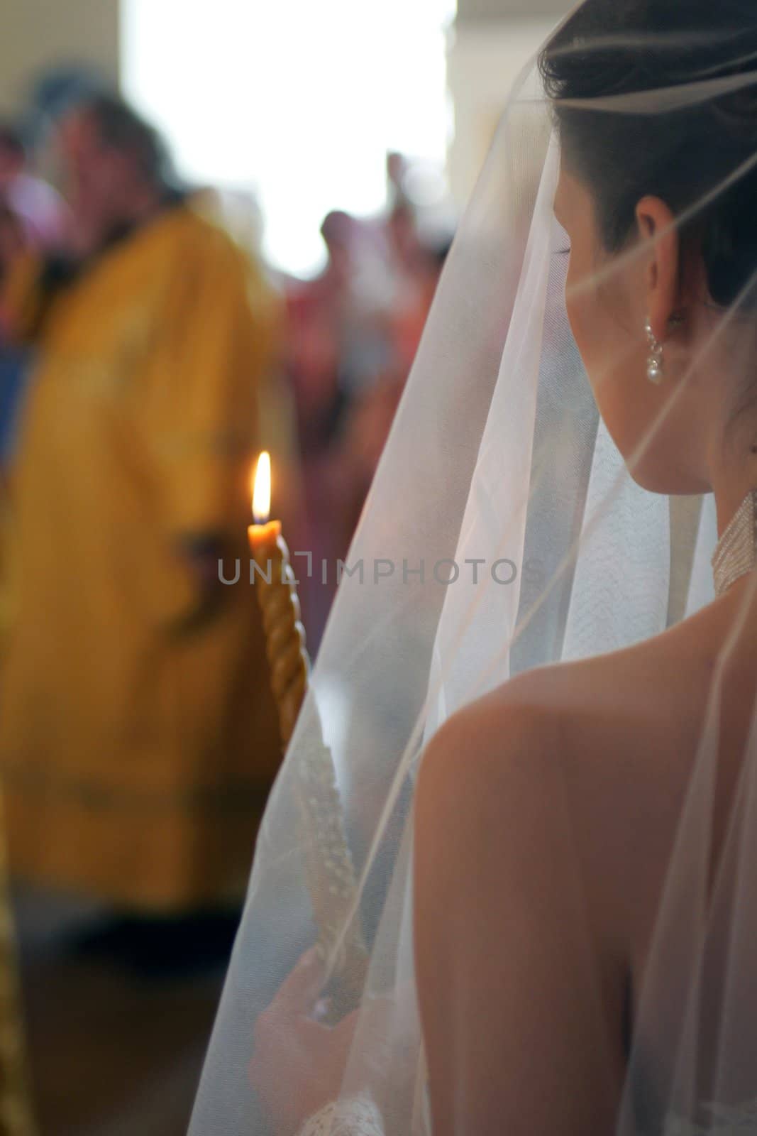 Rear view of bride wearing white wedding dress and holding lighted candle walking behind orthodox priest.
