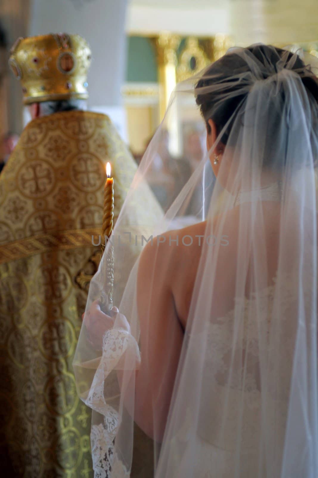 Rear view of bride wearing white wedding dress and holding lighted candle walking behind orthodox priest.