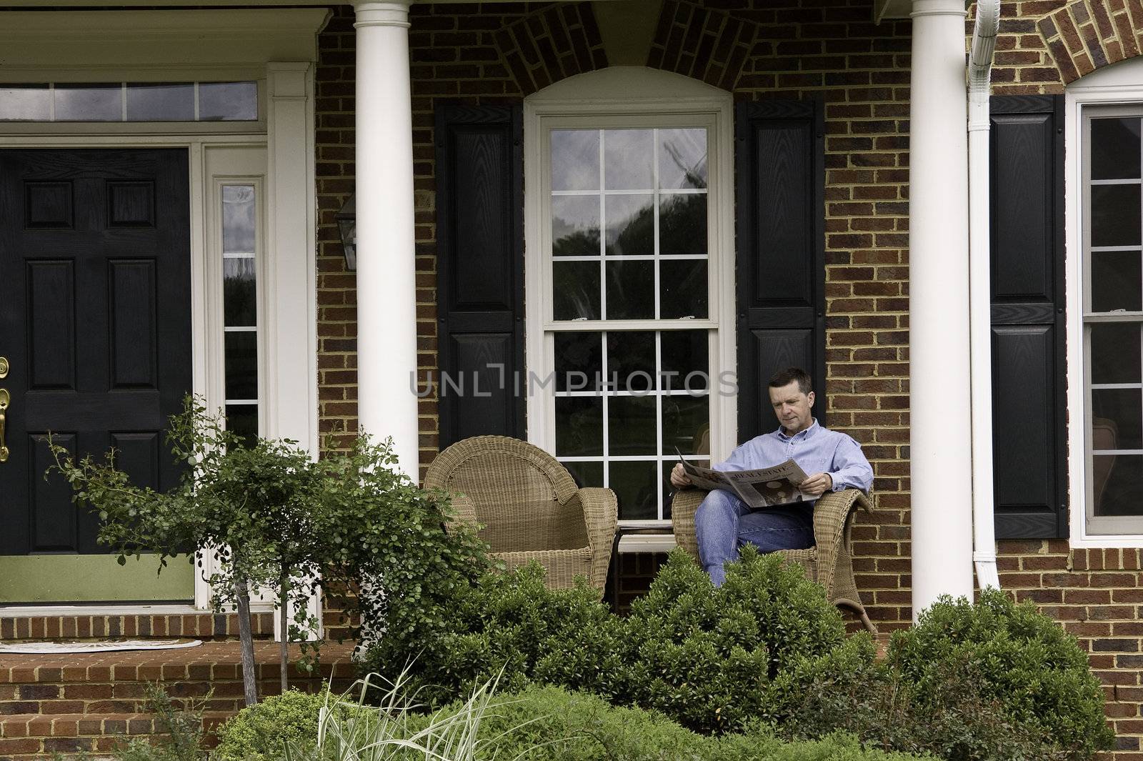 Middle aged man reading a newspaper on the porch of an expensive suburban house