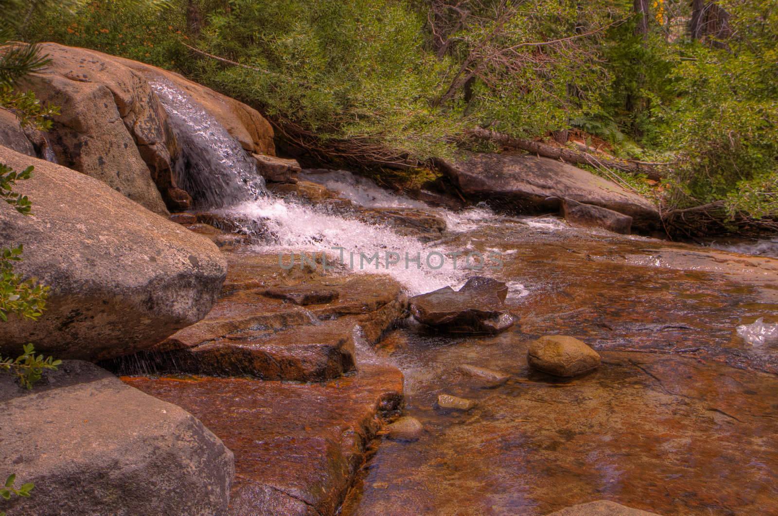 Small single waterfall cascading down mountain boulders into shallow water with forest background in HDR high dynamic range