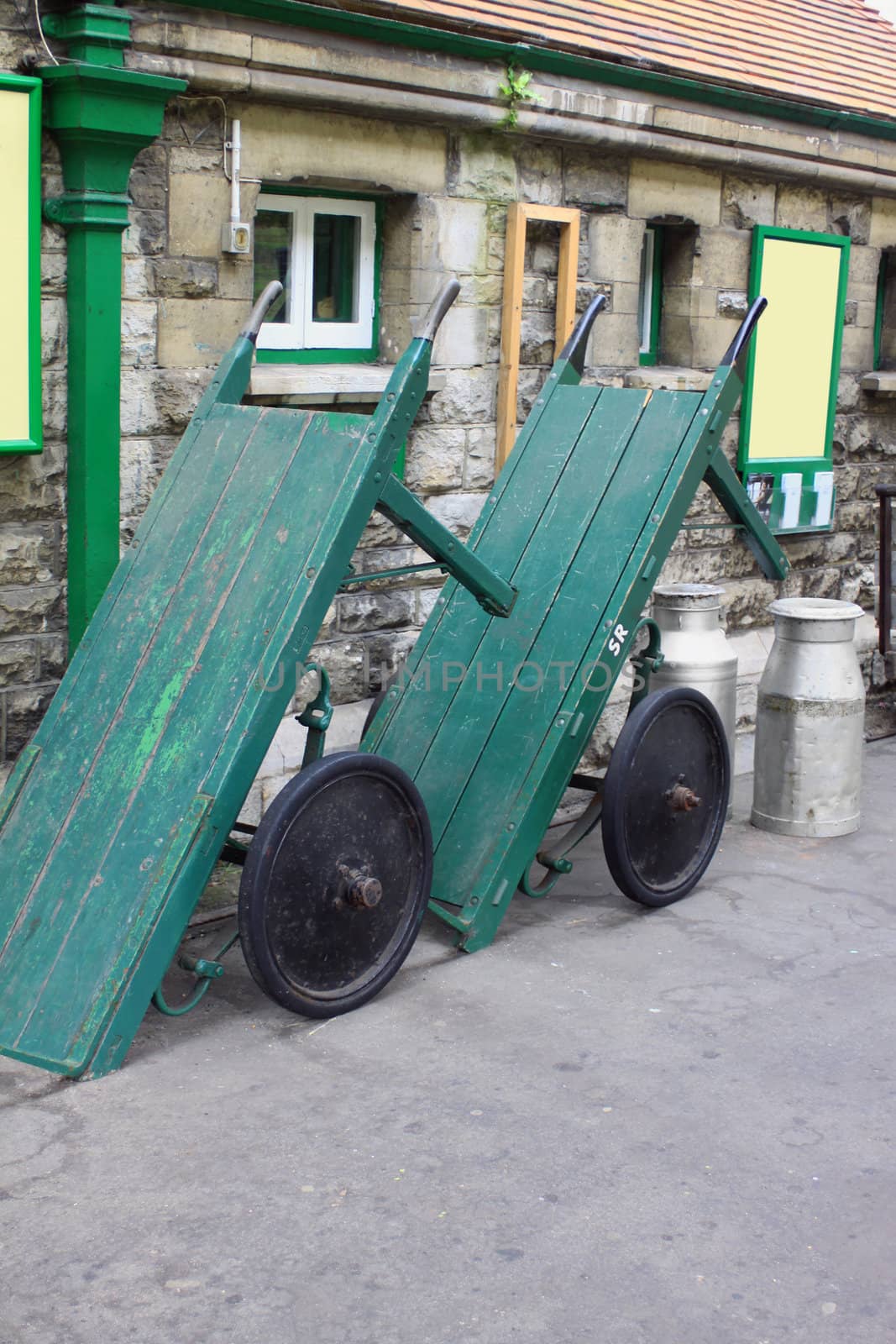 A pair of vintage wooden constructed baggage trollys on the platform of Swanage Railway in Dorset, UK. Set against a station building with two milk churns.