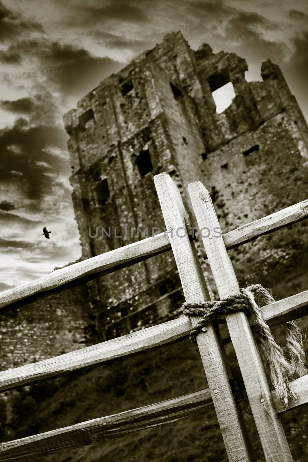 A mono image of Corfe Castle in Dorset with a dramatic cloud background and rolling fog at dusk, shot at a low angle with focus on a wooden gate to the foreground.