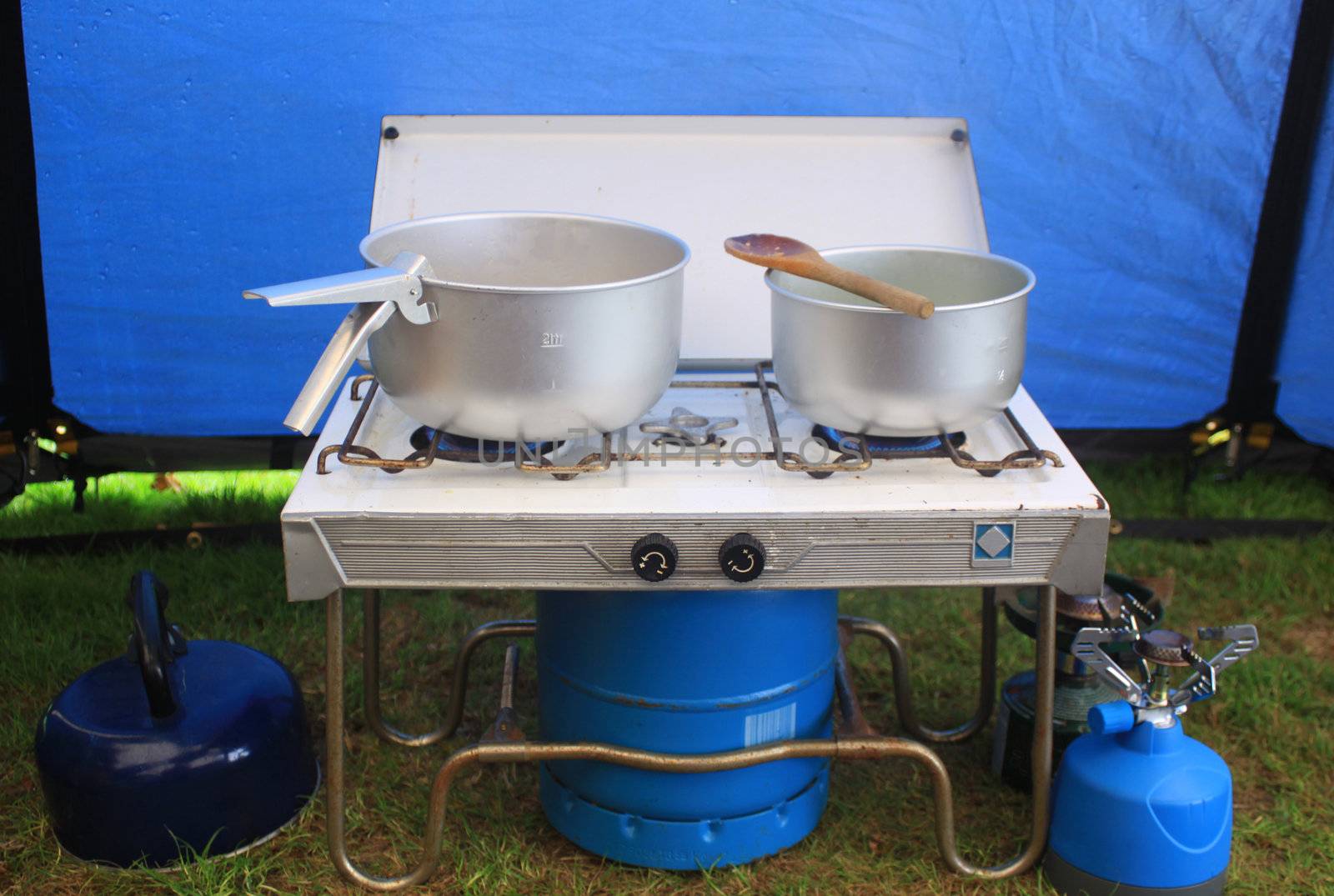 A two ringed gas camping stove. Set inside a blue canvas tent. Two tin pots are on the stove .
