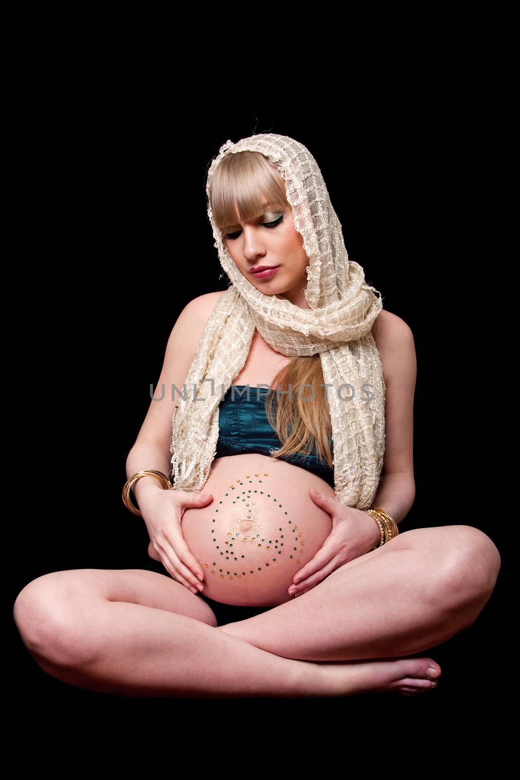 Beatiful pregnant Caucasian woman with head scarf sitting in lotus position holding naked belly covered with rhinestones, isolated