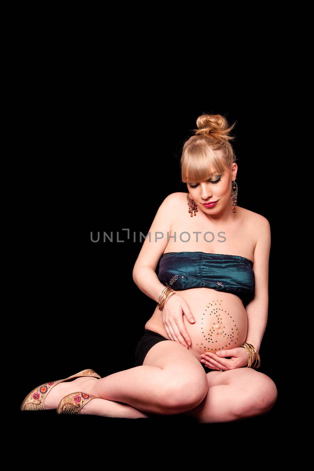 Beautiful pregnant blond girl holding her bare belly covered in rhinestones while looking down and sitting, isolated