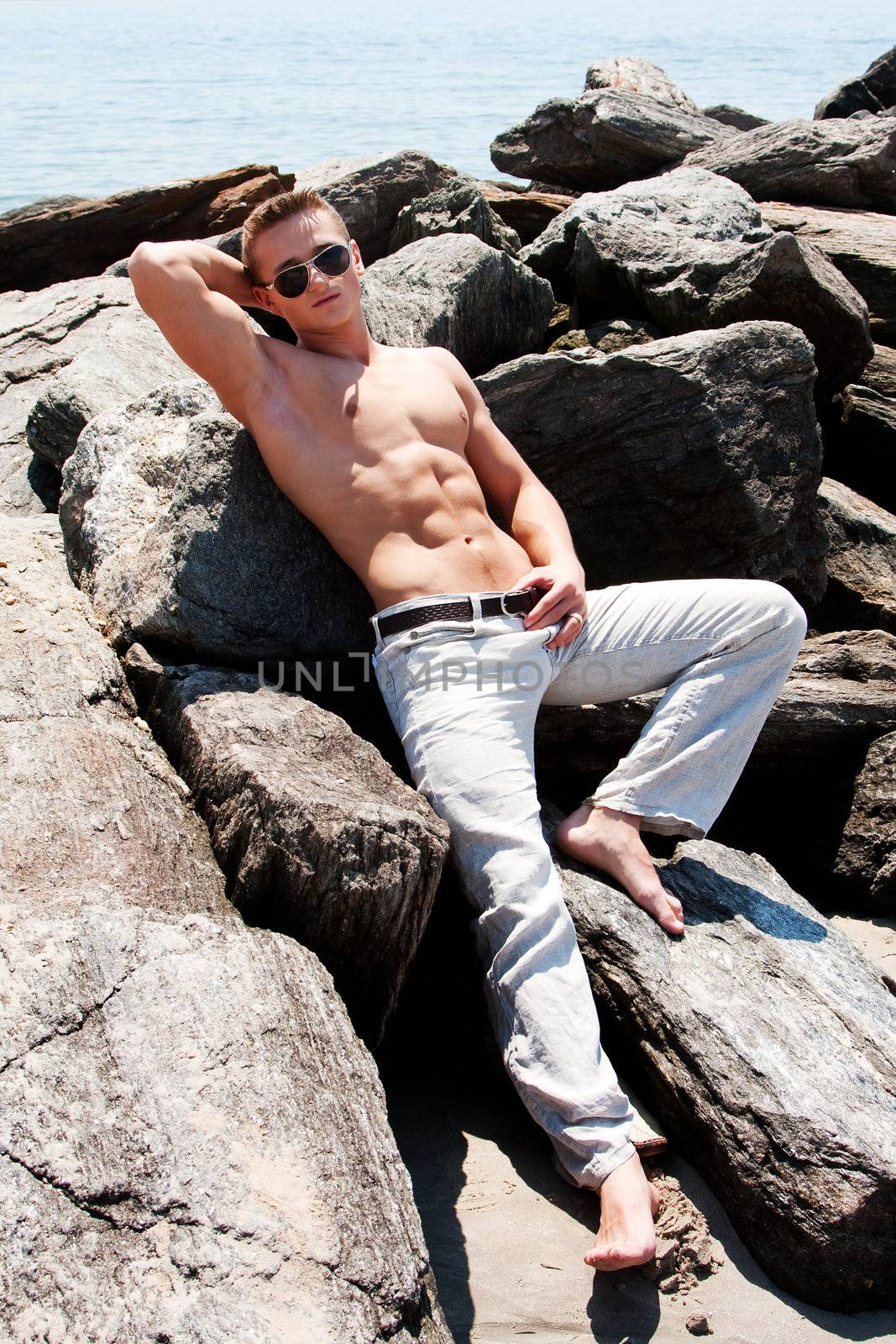 Sexy handsome male with sunglasses and toned body showing six pack abs on rocks at beach