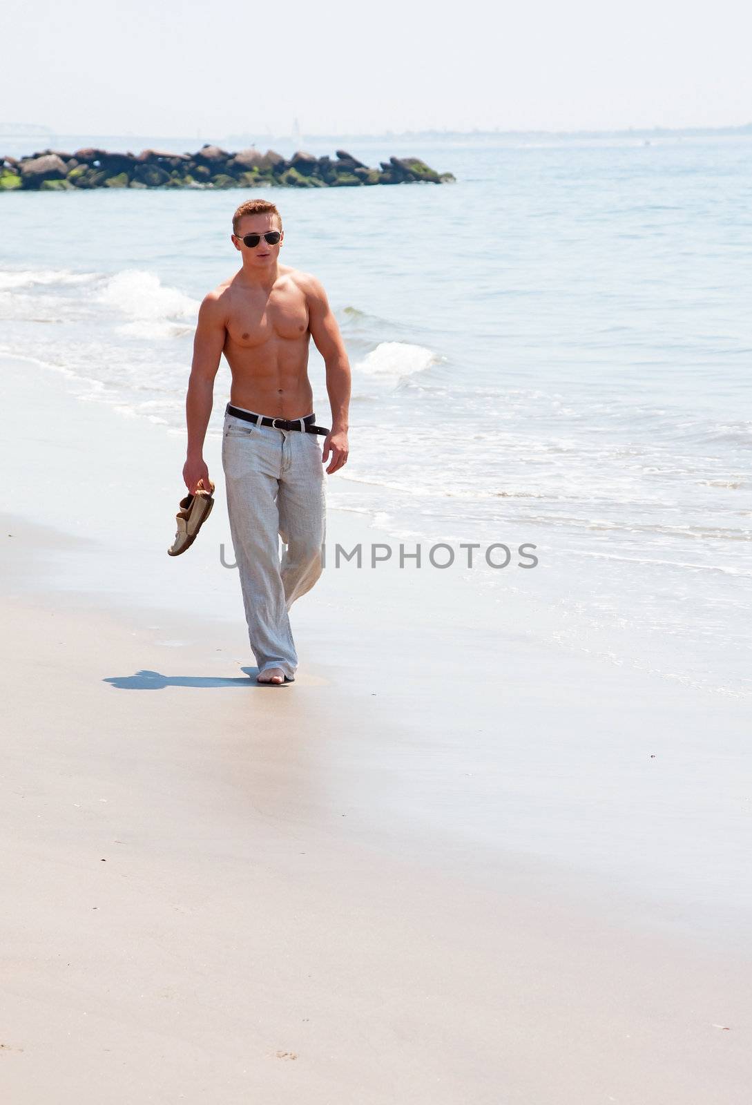 Toned cute handsome male walking on the beach with naked torso showing six pack abs holding shoes in hand wearing sunglasses