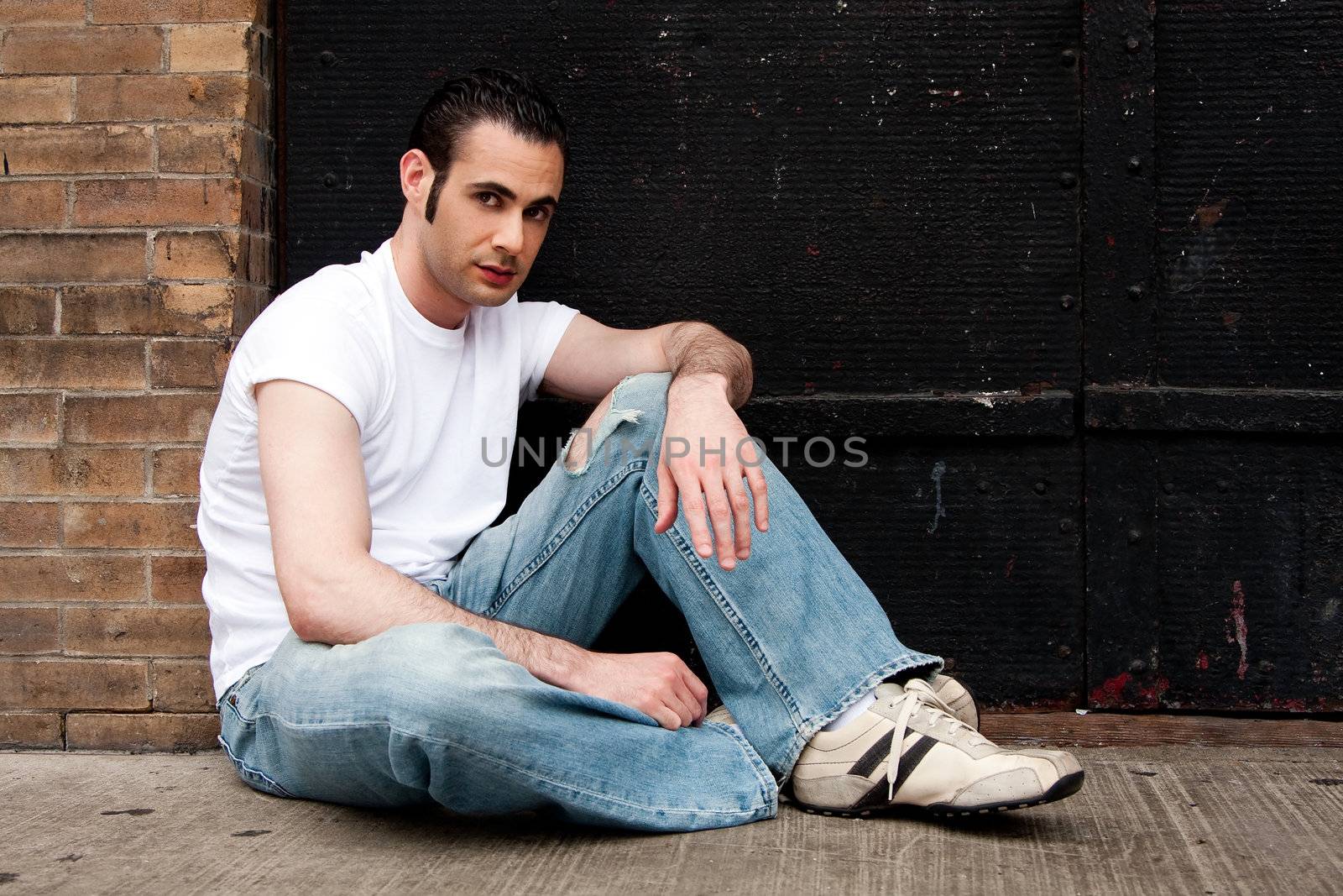 Handsome Caucasian man with sideburns dressed in white shirt and blue jeans sitting on concrete floor in front of black metal garage door