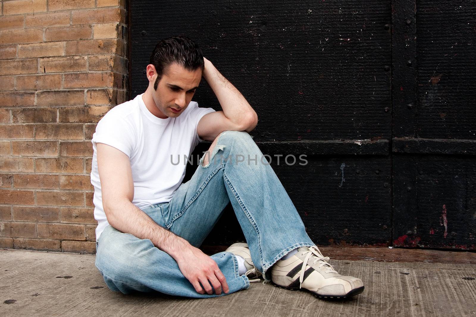 Handsome Caucasian man dressed in white shirt and blue jeans sitting on concrete floor in front of black metal door with a depressed expression thinking