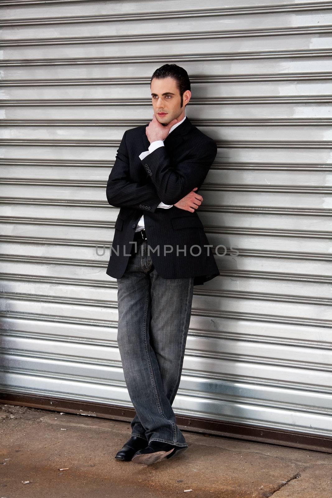 Handsome casual business man standing in front of and leaning against silver metal garage door with hand on chin thinking