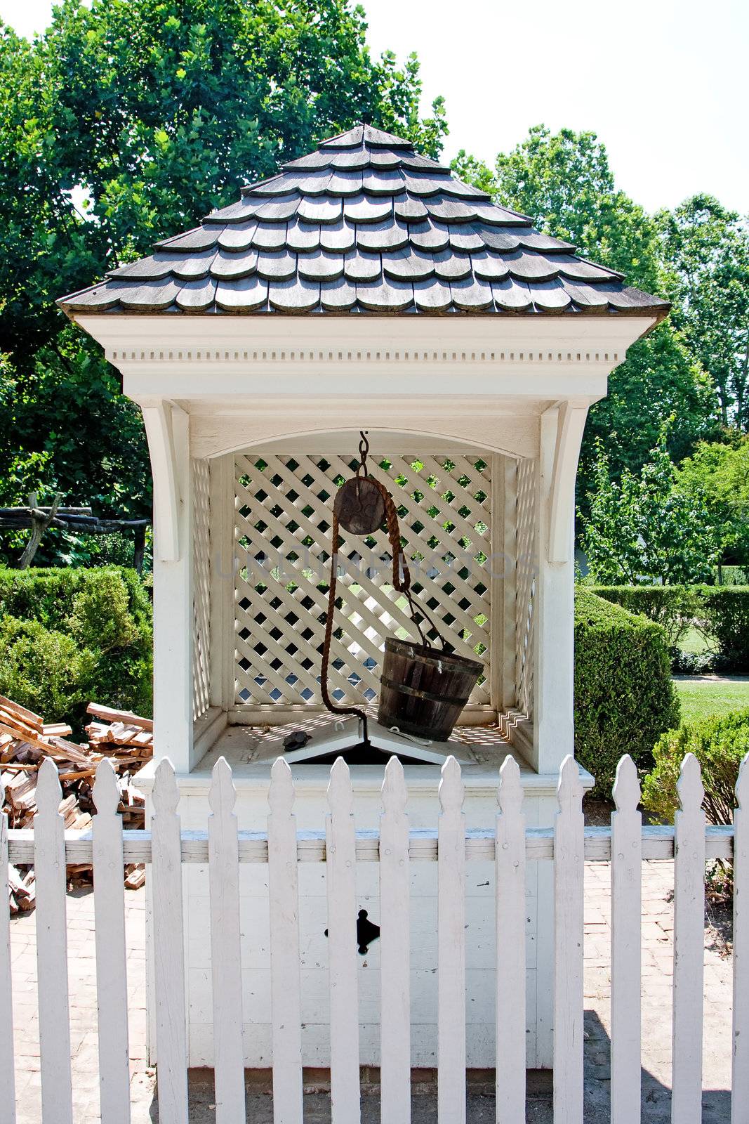 A white wishing water well in Colonial style with wooden bucket on top and roof with shingles behind a white picket fence in a garden.