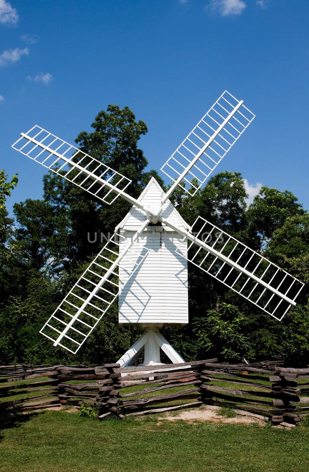 A white wooden windmill in a small meadow behind a wooden fence on a summer day with blue skies surrounded by trees.