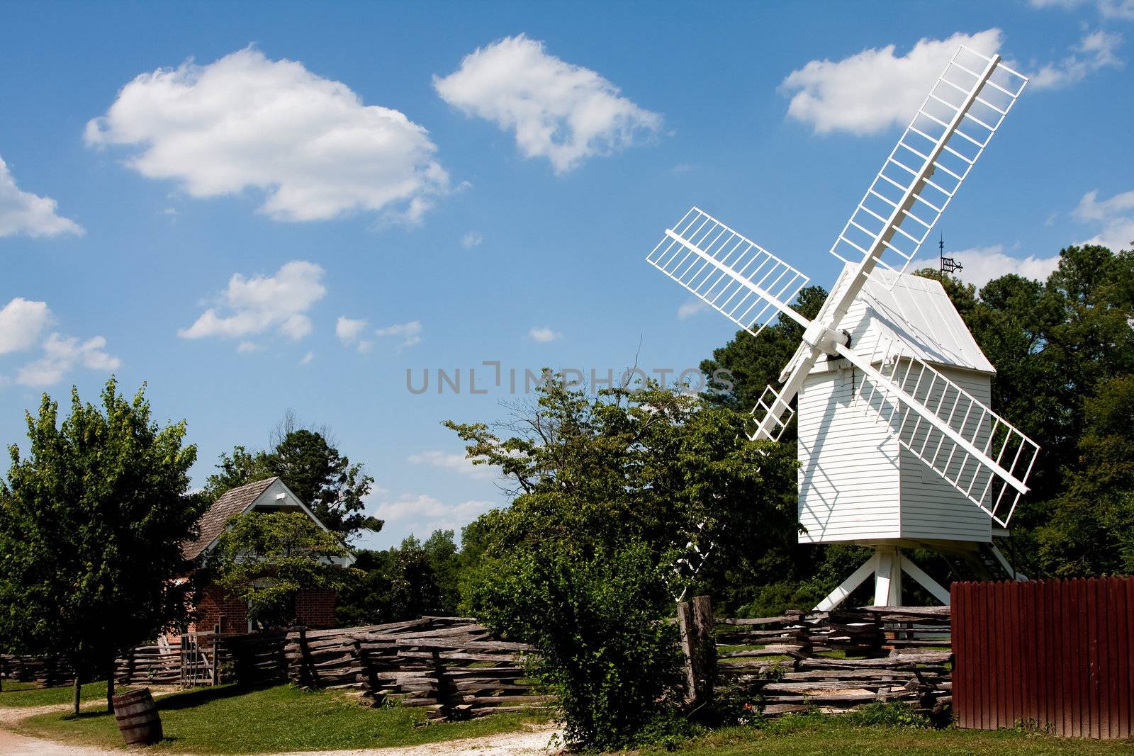 A white wooden windmill in a small meadow behind a wooden fence on a summer day with blue skies surrounded by trees in Colonial Williamsburg, Virginia.