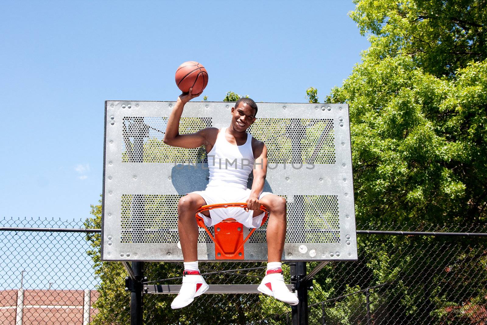 Happy smiling handsome sporty African-American male basketball player dressed in white and holding his ball with one hand outdoor on a summer day in a basketball court while sitting in the hoop.