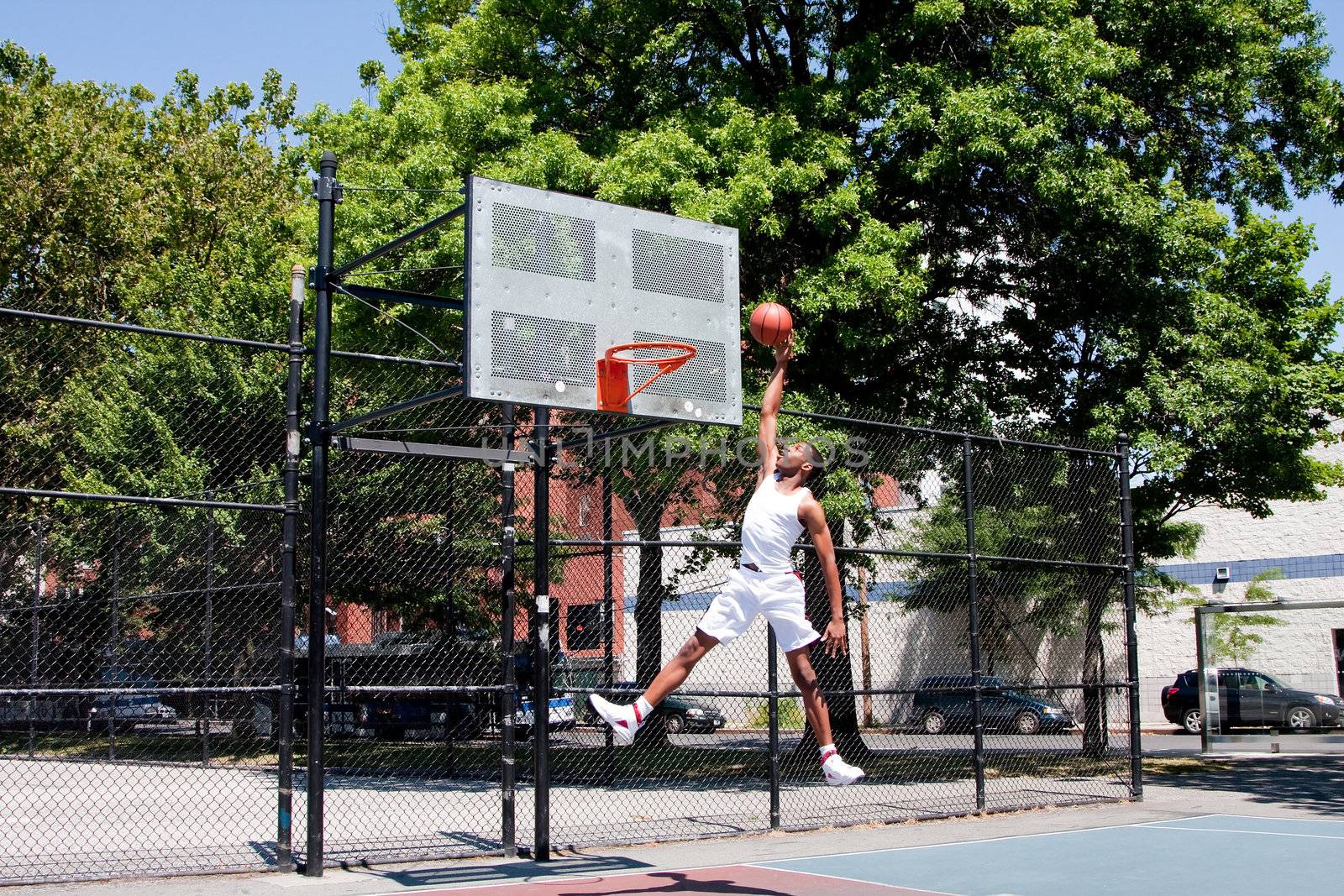 Sporty handsome African-American man dressed in white jumping in the air reaching for the basket while playing basketball in an outdoor court on a summer day