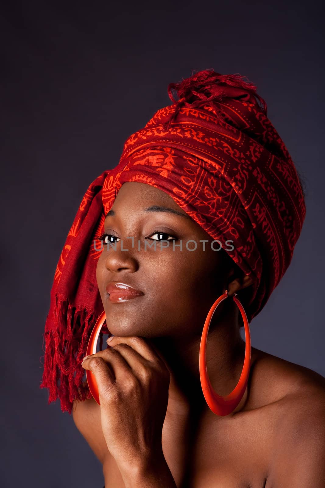 Beautiful African-American woman wearing a traditional tribal red orange head scarf and big orange hoop earrings, supporting her chin with hand, isolated.