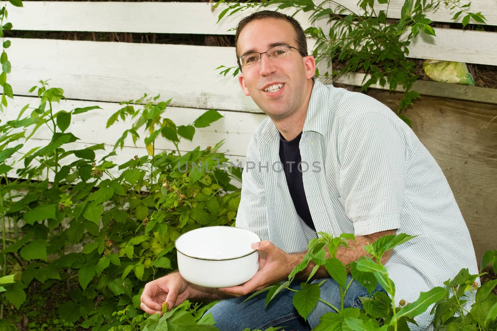 A young man wearing glasses is collecting raspberries of his garden outside
