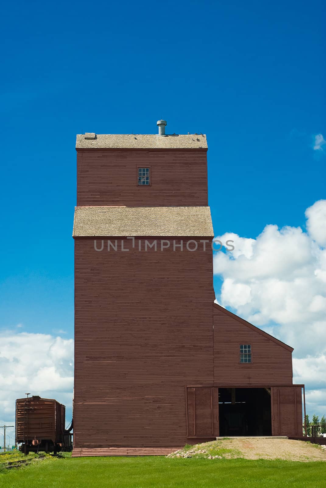 A Saskatchewan grain elevator with the bay doors open and a train parked beside it, shot on a partly cloudy day, with blue copy space above
