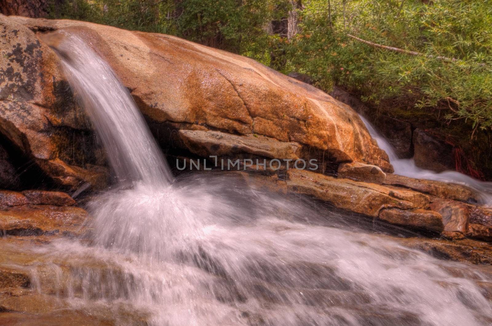 Small single waterfall cascading down mountain boulders into shallow water with forest background in HDR