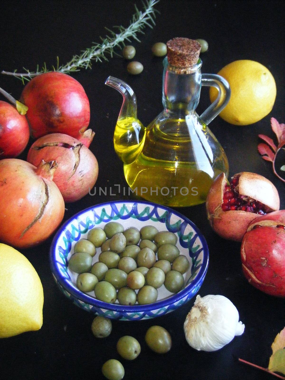 A jug of olive oil with olives and other fruits on a dark background.