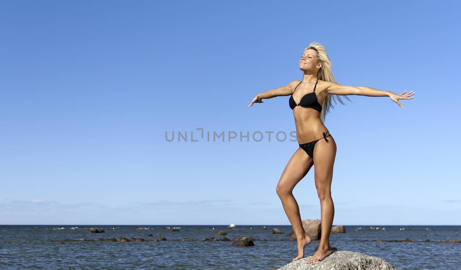 girl in bikini posing on a rock in a deep blue sea, arms outstretched to the sides