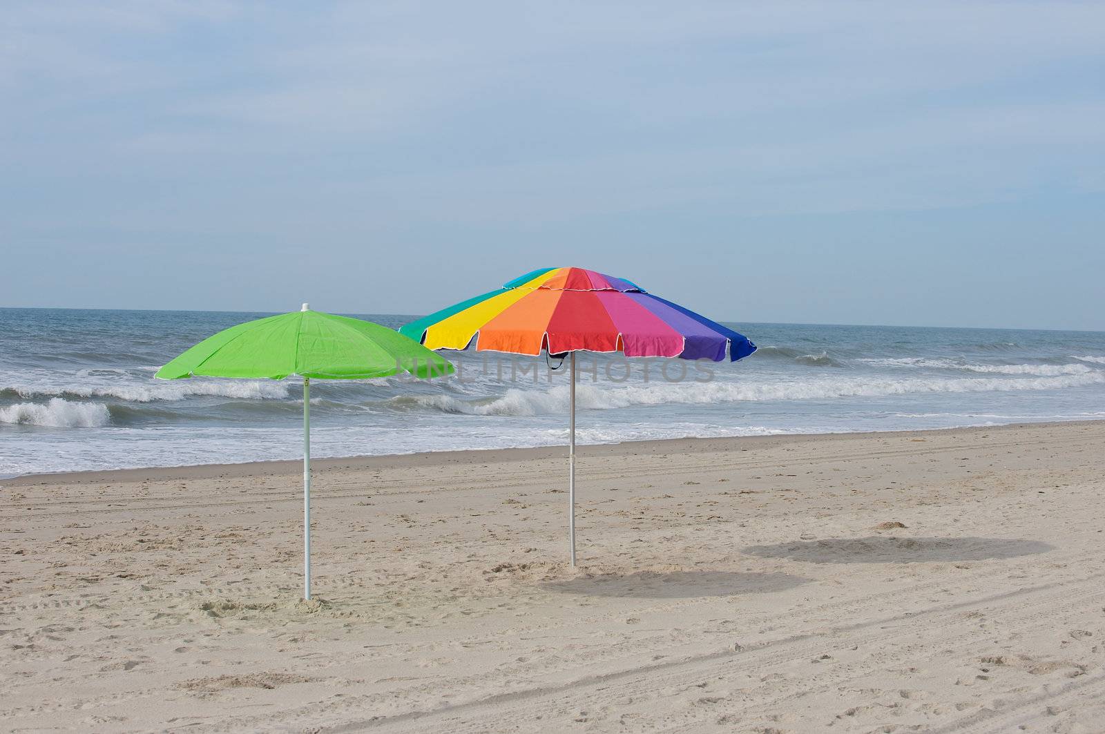 Two deserted umbrellas on the beach overlooking the surf, waiting for a tourist to claim them.