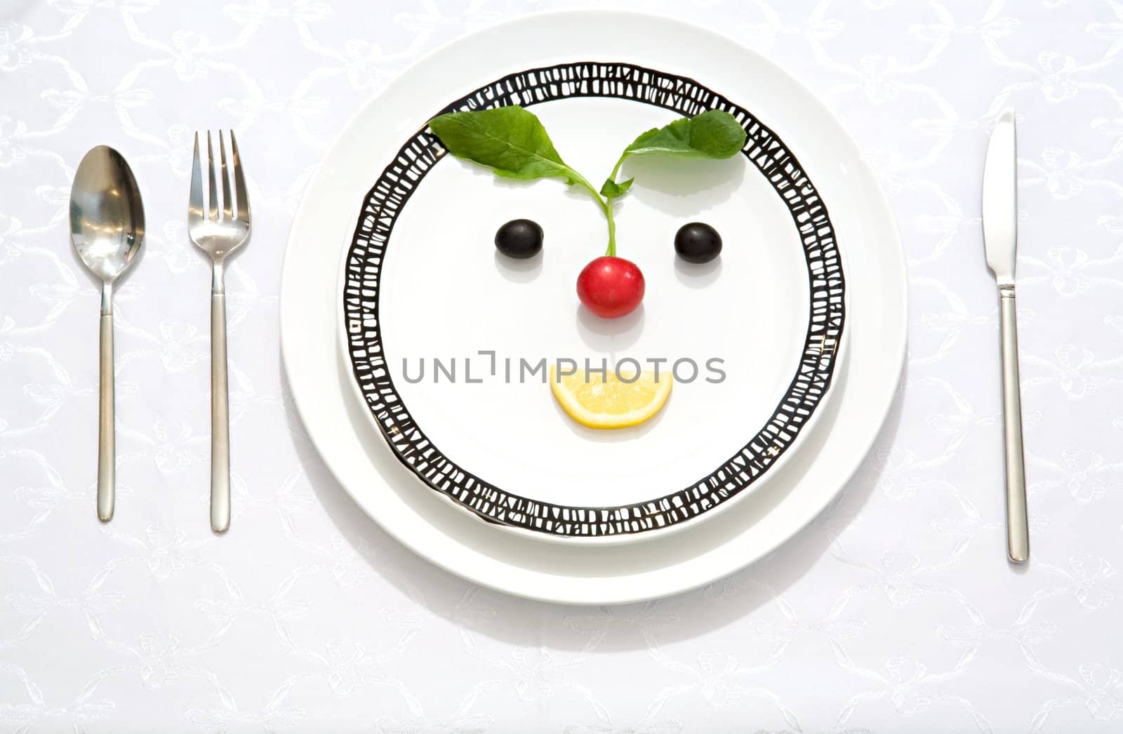The meal made as a smiling face from olives, radish and lemon slice on a flat plate with a knife, a fork and a spoon
