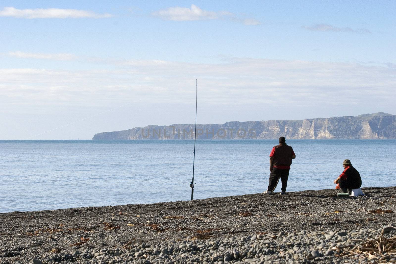 A couple of blokes sitting in the sun on an autumn day pretending to fish. Haumoana Beach, New Zealand. Cape Kidnappers in the background