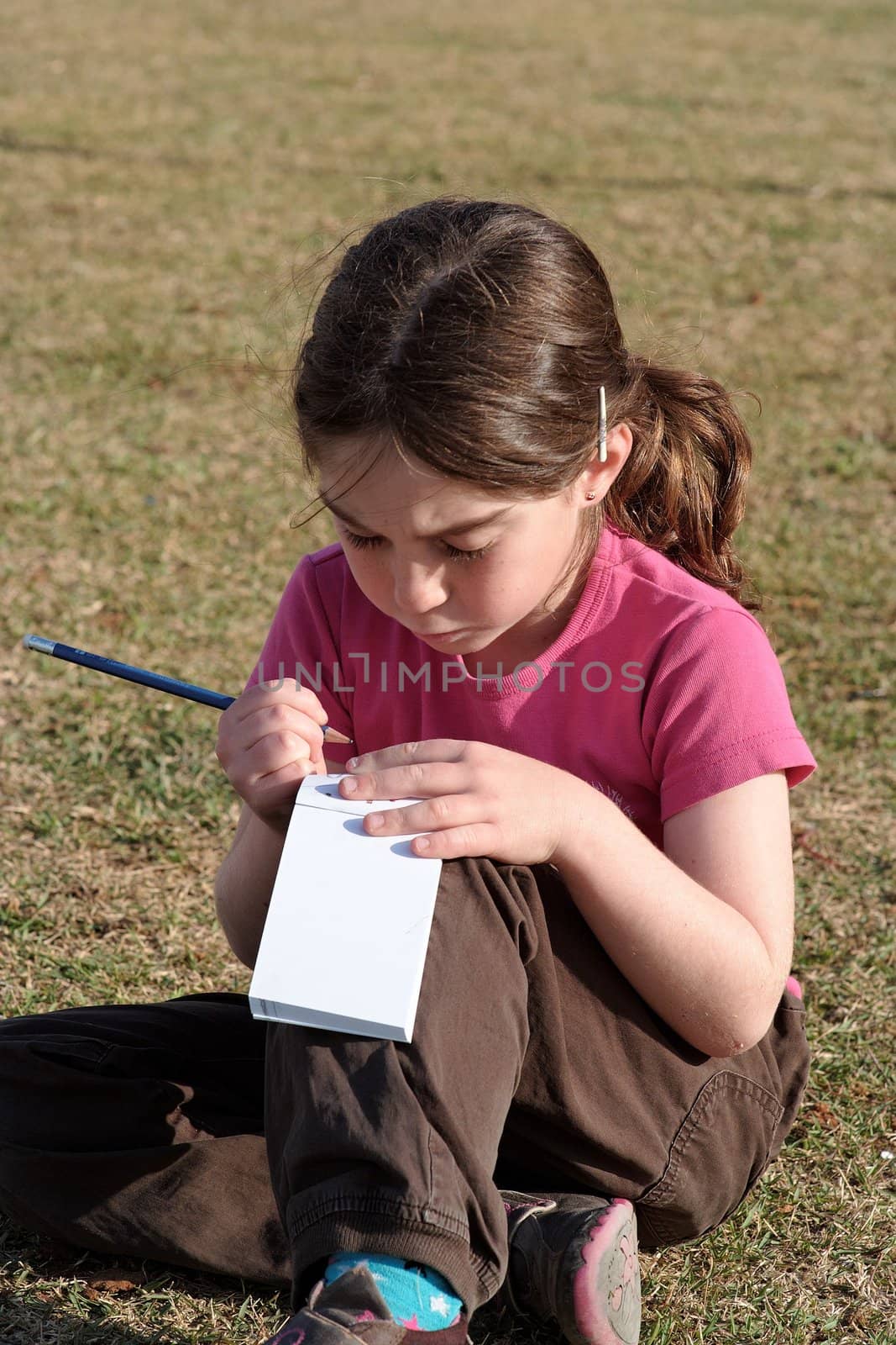 Cute little girl with pigtails writes in the notepad sitting on the grass outdoor