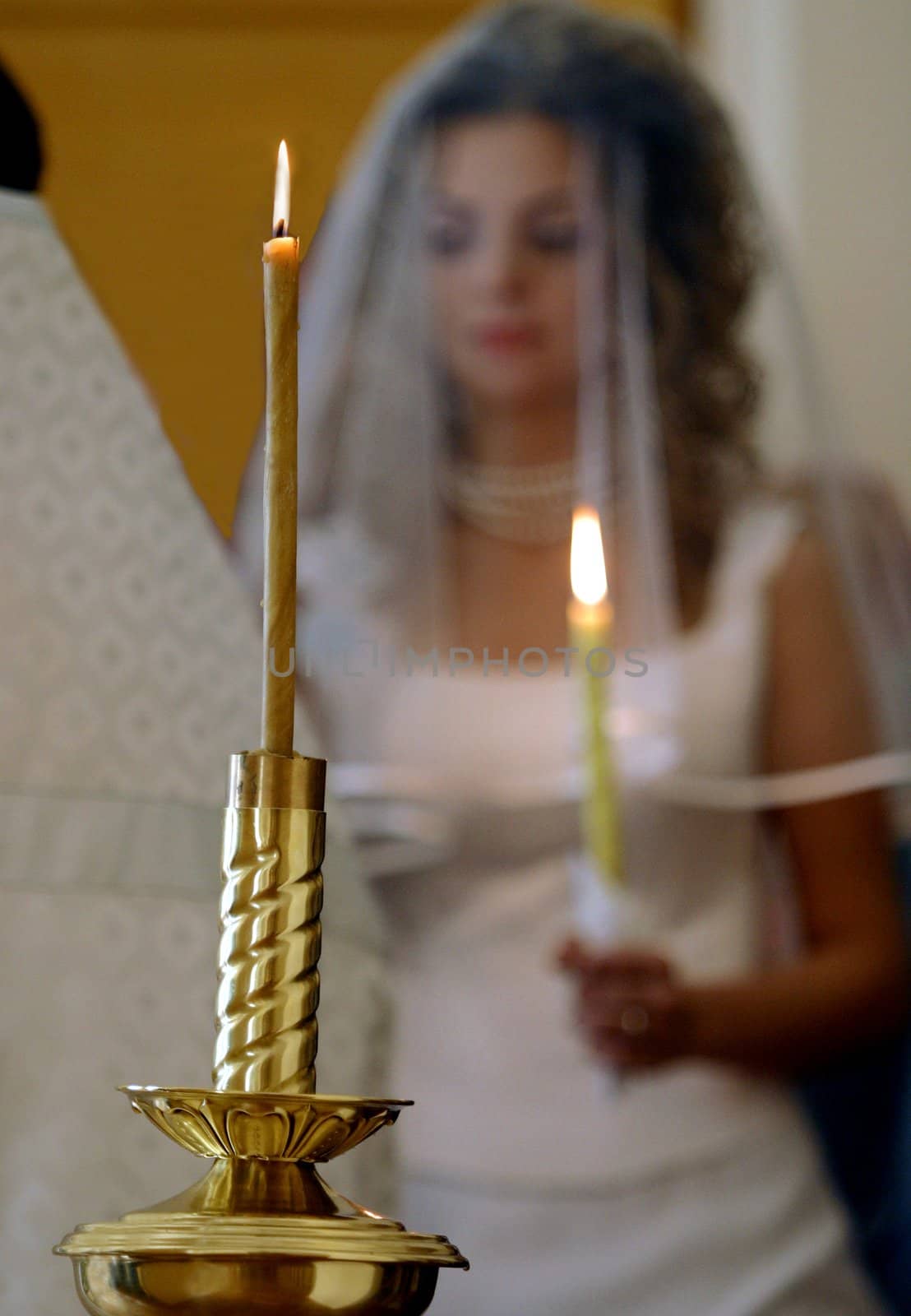 Bride wearing veil and holding lighted candle, golden candlestick in foreground - focus on candle.
