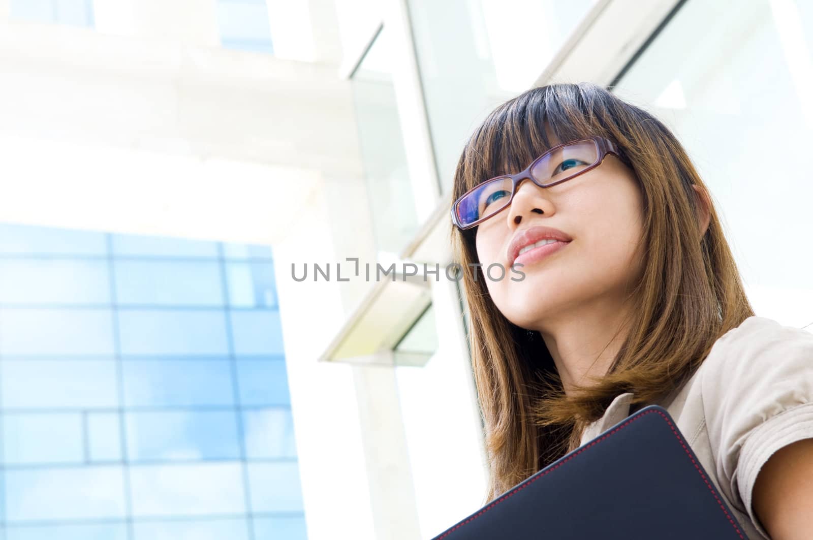 A young Asian woman looking far away to bright light in front of a modern office building, with diary on hand.