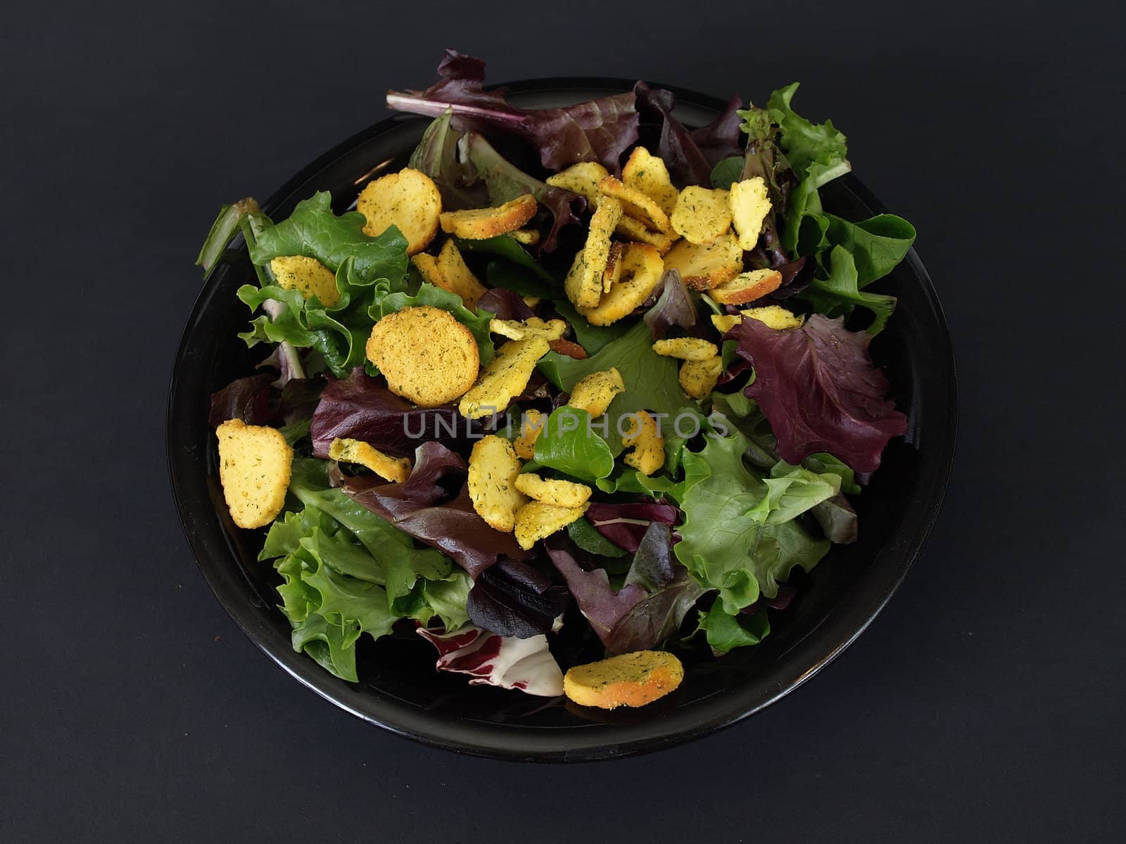 A black plate of salad with crutons and green and purple leaves over a black background.