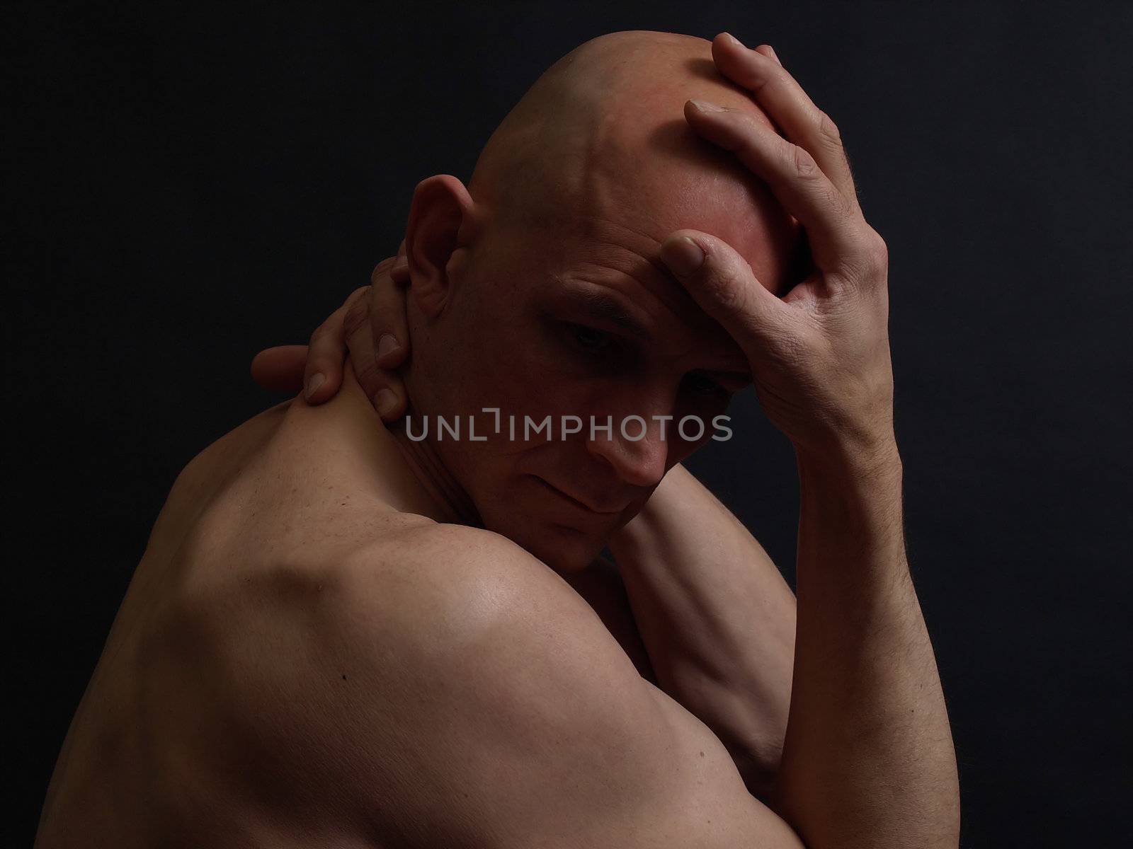 A nude bald male in the shadows over a black background.