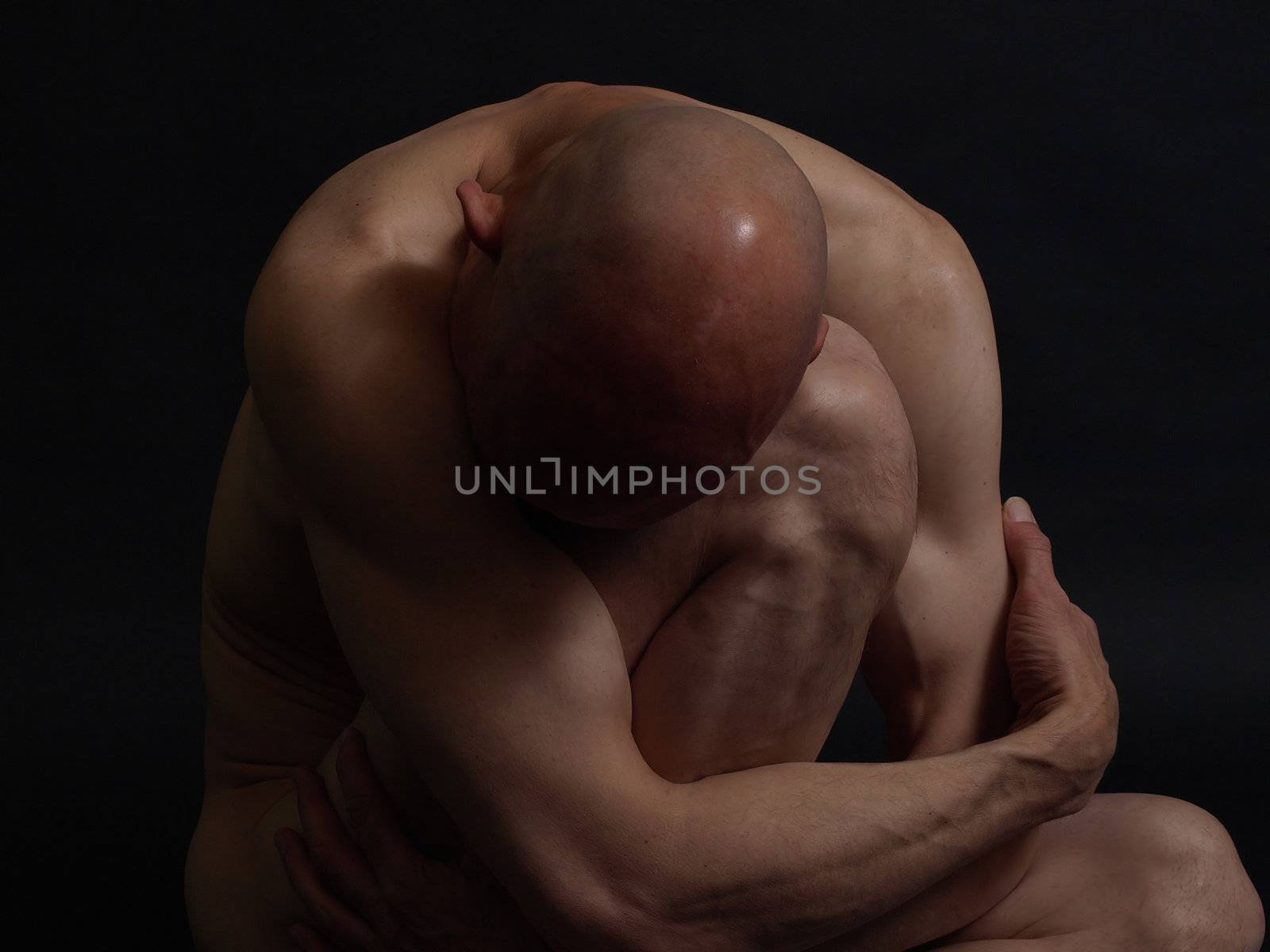 A clean shaven nude male sits in darkness, crumpled up. Over a black background.