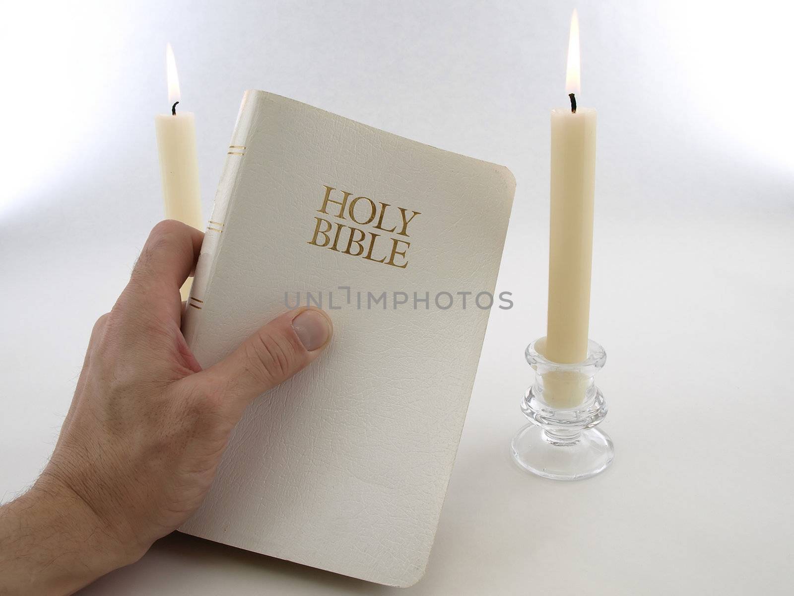A man's hand holds up a white Holy Bible. Two taper candles burn in the background. Over a white background.