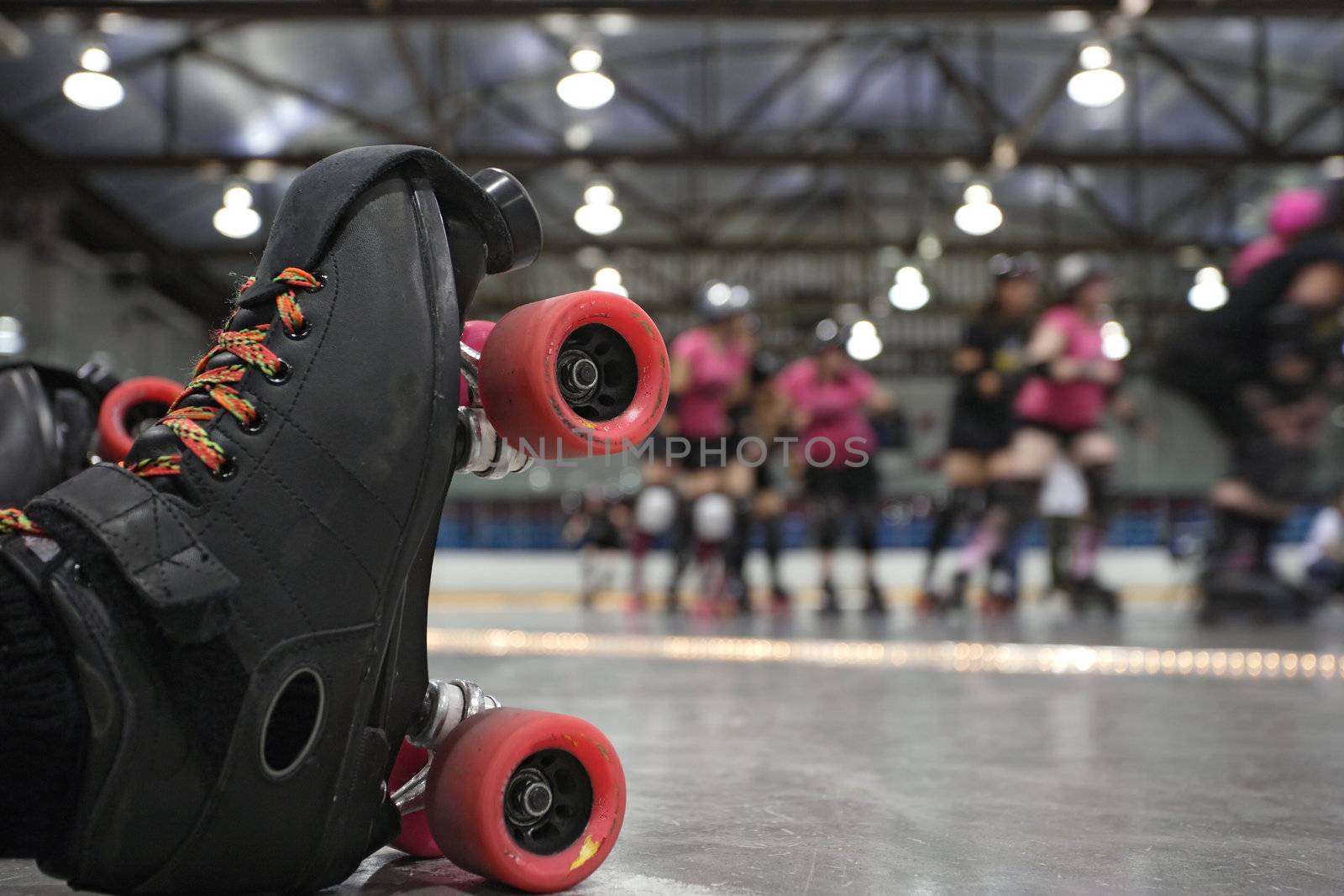 An abstract image of the roller-skates of a fallen skater as her teammates in the background continue to skate around the track of the roller derby.
