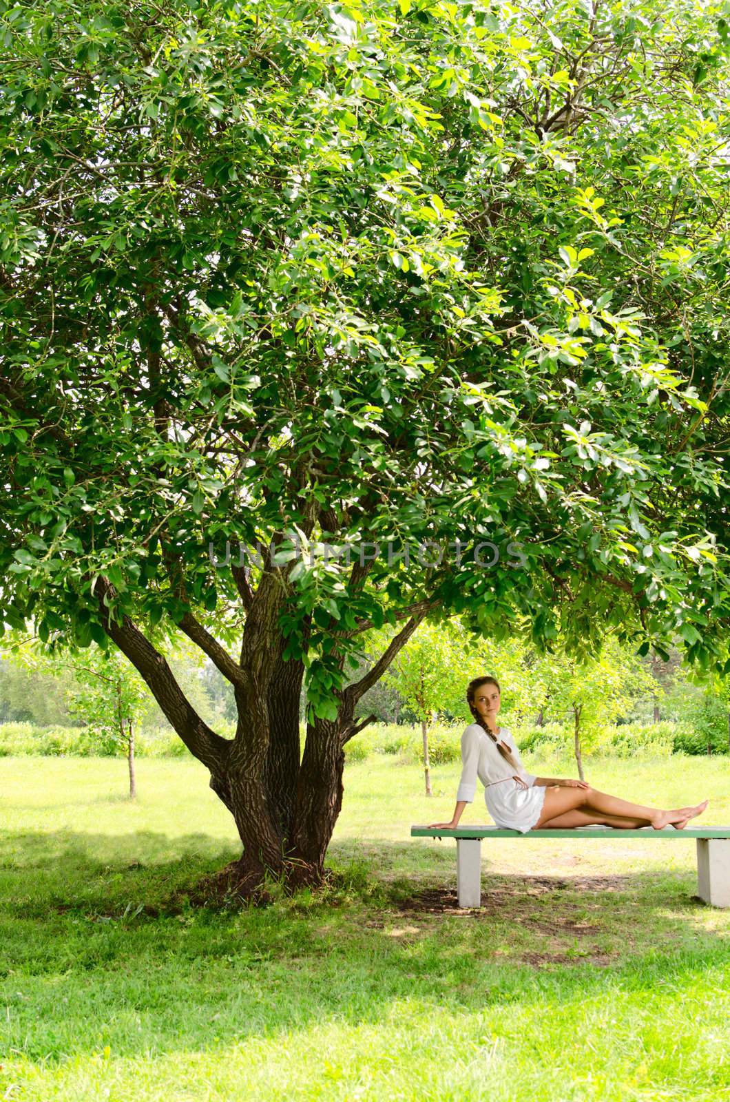 Pretty barefooted woman is sitting on a bench under the green tree. 
