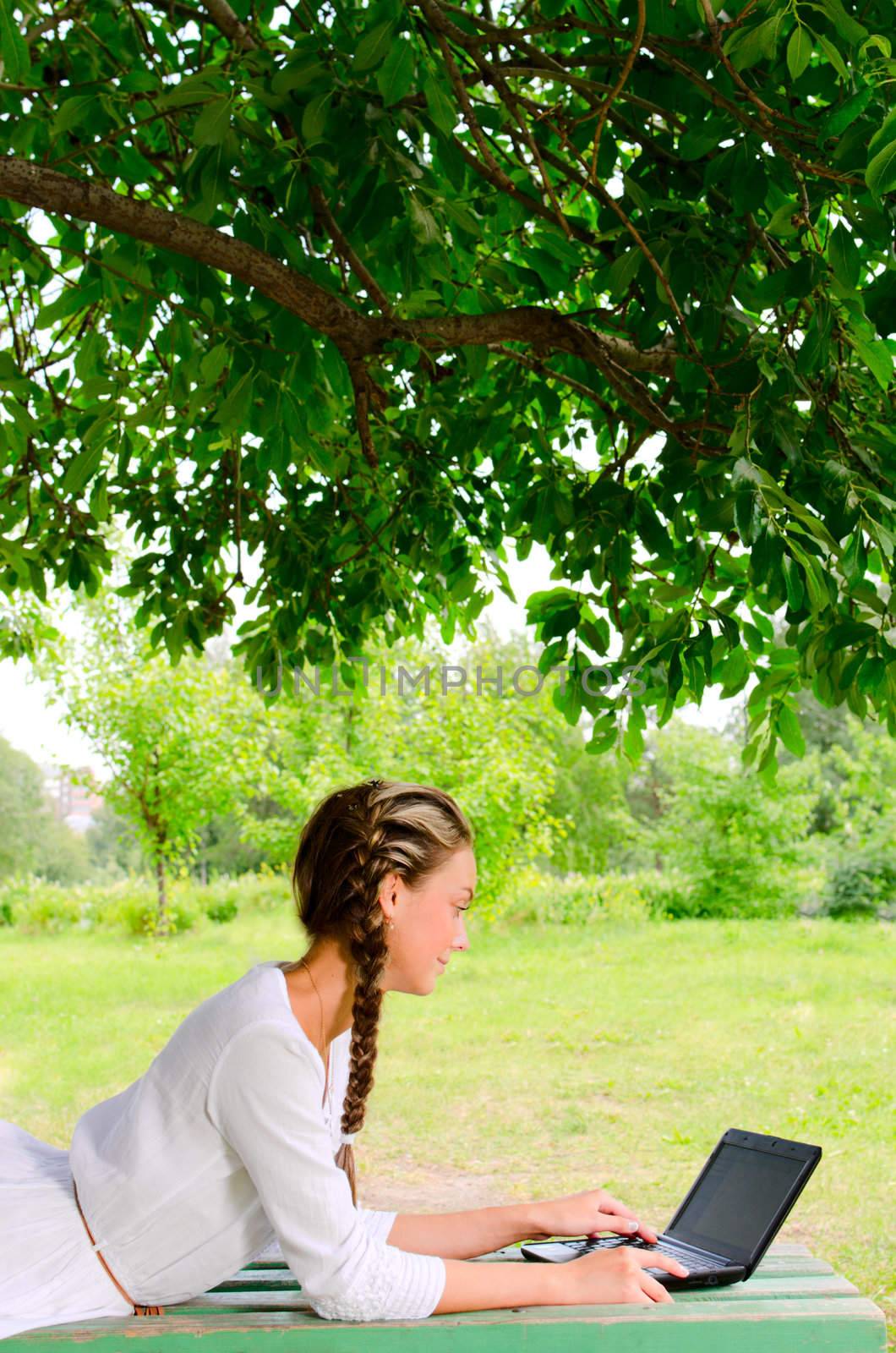 Pretty barefooted woman is laying on a bench under the green tree. She is working with small notebook