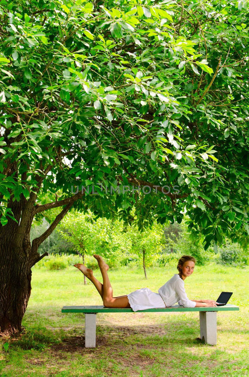 Pretty barefooted woman is laying on a bench under the green tree. She is working with small notebook. She is looking at the camera and slightly smiling