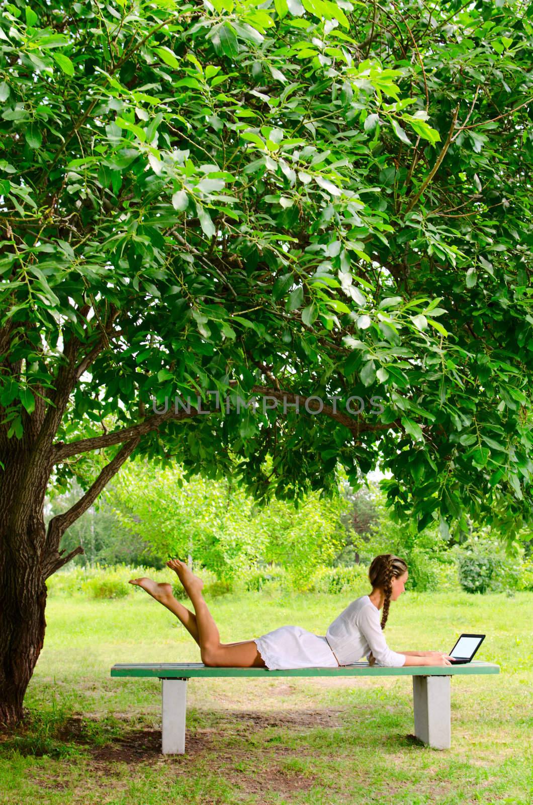 Pretty barefooted woman is laying on a bench under the green tree. She is working with small notebook